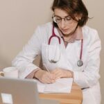 Female doctor with stethoscope writing notes in an office setting with laptop.